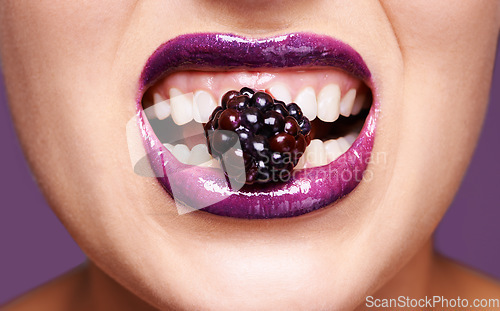 Image of Woman lips, purple lipstick with berry and makeup, shine and creativity with beauty isolated on studio background. Closeup of fruit between female model teeth, cosmetic product and cosmetology