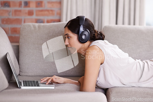 Image of Woman, laptop and lying on sofa with headphones for streaming, music or search for web video in home. Girl, computer or scrolling for movie, podcast or audio on internet to relax on living room couch