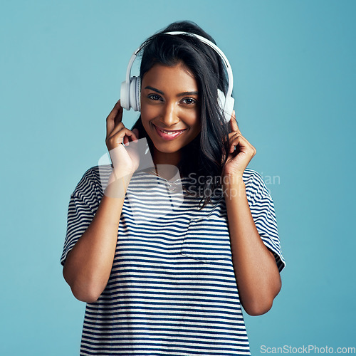 Image of Woman, portrait and headphones with music in studio with happiness and web audio. Blue background, Indian female person and young model listening, hearing and streaming a song with smile and online