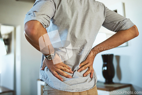 Image of Hands, back pain and injury with a man standing in the living room of his home, holding his spine in discomfort. Health, medical and anatomy with a male person in a house feeling a muscle cramp