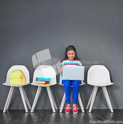 Image of Child, chair and laptop for learning and education while typing for research on internet. Happy girl kid or student against a grey wall with technology, book and backpack in a waiting room with space