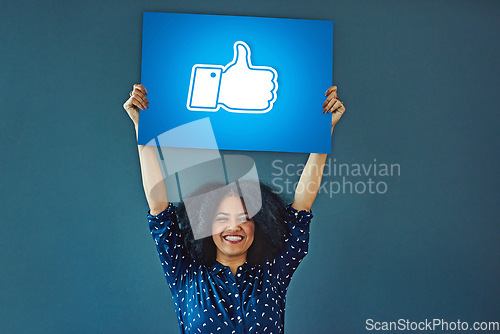 Image of Portrait, social media and thumbs up icon to like with a woman holding a poster in studio on a blue background. Smile, logo and button with a happy female influencer showing a hand gesture sign