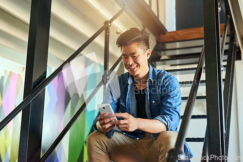 Image of Asian man, texting and office stairs with smartphone, smile and communication on social network app. Young japanese creative, web design startup and relax on steps in workplace for chat with phone