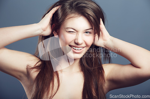 Image of Wink, face and beauty portrait of a woman in studio with a smile, makeup and hands in hair. Headshot of a happy female model on a grey background for natural glow, skin cosmetics and playful flirting