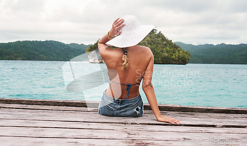 Image of Travel, hat and back of woman on an island to relax while on tropical summer vacation or holiday. Adventure, outdoor and female person sitting on wood pier by ocean water on weekend trip in paradise.