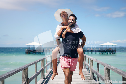 Image of Happy couple, vacation and walking on ocean boardwalk for piggy back, freedom and travel. A man and woman playing and laughing on tropical walk to relax on holiday, nature adventure or date at sea