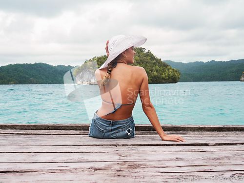 Image of Adventure, hat and woman on an island to relax while on tropical summer vacation or holiday. Travel, outdoor and back of female person sitting on wood pier by ocean water on weekend trip in paradise.