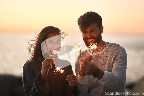 Image of Sunset, beach and couple with fireworks for celebration, party and sparks on romantic date. Nature, love and happy man and woman with sparklers for honeymoon on holiday, weekend and vacation