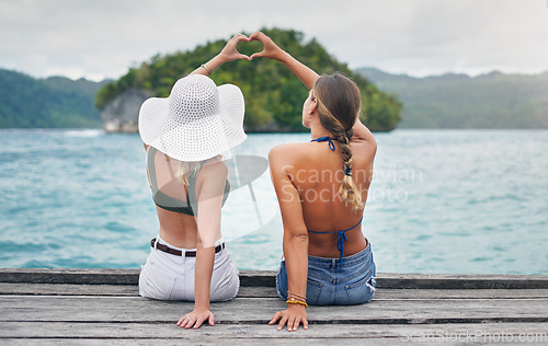 Image of Heart hands, back and girl friends by a lake while on a summer, paradise and tropical vacation. River, dock and women sitting to relax on wood pier with love shape gesture on holiday or weekend trip.