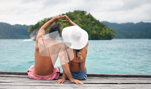 Image of Heart hands, back and couple by a ocean while on summer, paradise and tropical vacation. Romance, dock and young man and woman relaxing on wood pier with love shape gesture on holiday or weekend trip