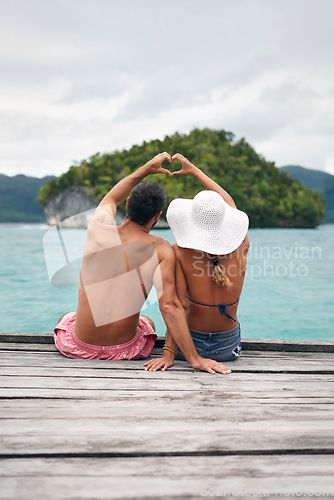 Image of Heart shape, hands and couple by ocean while on a summer, romantic and tropical vacation. Island, dock and back of man and woman relaxing on wood pier with love gesture on holiday or weekend trip.