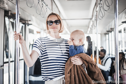 Image of Mother carries her child while standing and holding on to bar holder on bus. Mom holding infant baby boy in her arms while riding in public transportation. Cute toddler traveling with mother.