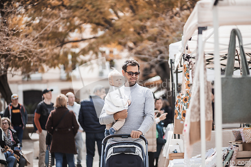 Image of Father walking carrying his infant baby boy child and pushing stroller in crowd of people wisiting sunday flea market in Malaga, Spain