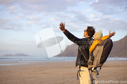 Image of Young father rising hands to the sky while enjoying pure nature carrying his infant baby boy son in backpack on windy sandy beach of Famara, Lanzarote island, Spain at sunset. Family travel concept.