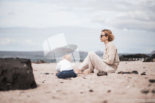 Image of Mother enjoying winter beach vacations playing with his infant baby boy son on wild volcanic sandy beach on Lanzarote island, Canary Islands, Spain. Family travel and vacations concept