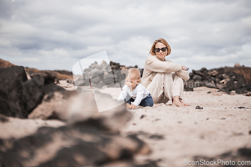 Image of Mother enjoying winter beach vacations playing with his infant baby boy son on wild volcanic sandy beach on Lanzarote island, Canary Islands, Spain. Family travel and vacations concept