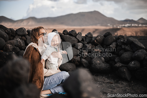 Image of Mother enjoying winter vacations playing with his infant baby boy son on black sandy volcanic beach of Janubio on Lanzarote island, Spain on windy overcast day. Family travel vacations concept.