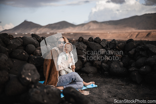 Image of Mother enjoying winter vacations playing with his infant baby boy son on black sandy volcanic beach of Janubio on Lanzarote island, Spain on windy overcast day. Family travel vacations concept.