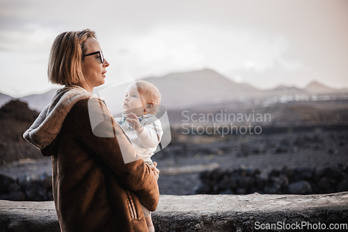 Image of Mother enjoying winter vacations playing with his infant baby boy son on black sandy volcanic beach of Janubio on Lanzarote island, Spain on windy overcast day. Family travel vacations concept.