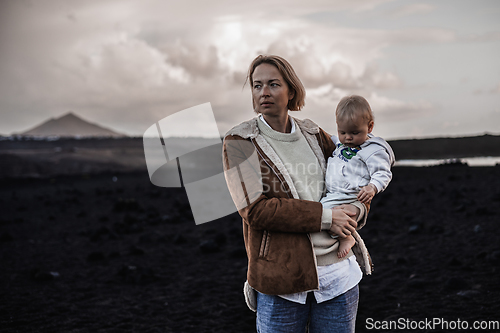 Image of Mother enjoying winter vacations playing with his infant baby boy son on black sandy volcanic beach of Janubio on Lanzarote island, Spain on windy overcast day. Family travel vacations concept.