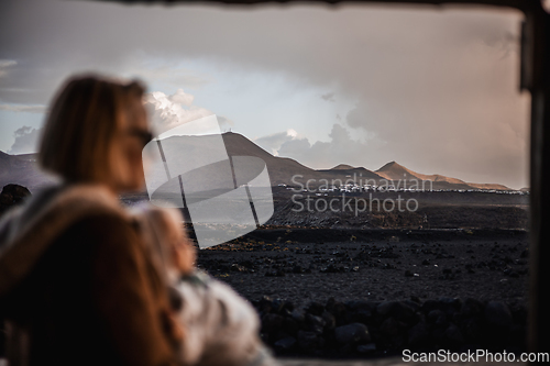 Image of Mother enjoying winter vacations playing with his infant baby boy son on black sandy volcanic beach of Janubio on Lanzarote island, Spain on windy overcast day. Focus on landscape in background.