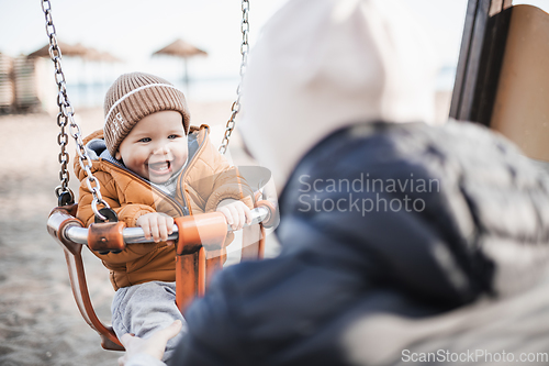 Image of Mother pushing her cheerful infant baby boy child on a swing on sandy beach playground outdoors on nice sunny cold winter day in Malaga, Spain.