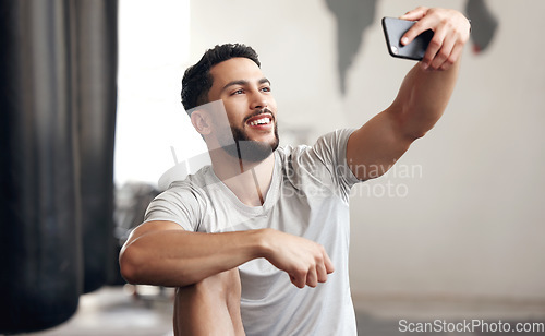 Image of One fit young hispanic man using a cellphone to take selfies while on a break from exercise in a gym. Happy mixed race guy making video call and taking photos for social media during a rest from trai