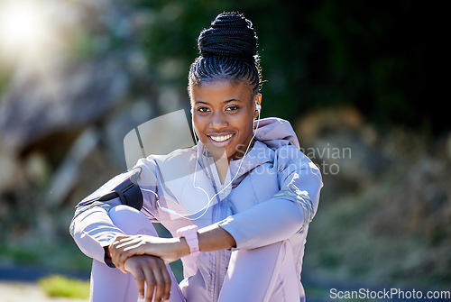 Image of Fitness, portrait and woman resting after a workout in nature with earphones for music, radio or podcast. Happy, smile and African female athlete sitting after an outdoor cardio running exercise.