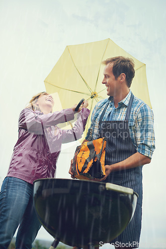 Image of Man, woman and outdoor for bbq in rain with umbrella, charcoal and surprise on vacation in low angle. Couple, outdoor and grill in storm, cooking and insurance in winter with comic laughing in garden