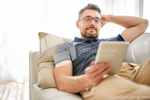 Image of Tablet, relax and man on a sofa in the living room watching a video on social media or the internet. Rest, browsing and mature male person reading an online blog on digital technology at his home.