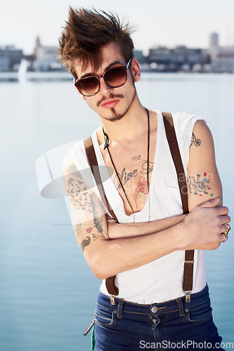 Image of Sunglasses, portrait and man by sea with arms crossed for stylish tattoo, body art and fashion. Hipster, ocean and confident male person standing outdoor in Spain with cool clothes, attitude and punk