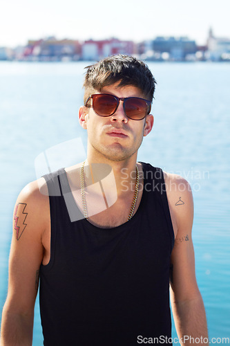 Image of Portrait, sunglasses and serious man by ocean with stylish tattoo, body art and fashion. Face, sea and trendy male person standing outdoor in Norway with cool clothes, attitude and youth aesthetic.