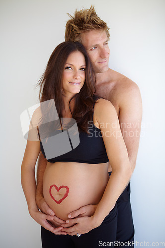 Image of Pregnant, hug and portrait of couple with heart on stomach for love, care and support on white background. Family, pregnancy and man and woman in studio excited for baby with love symbol on belly