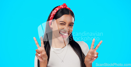 Image of Face, funny and Indian woman with peace sign, confident and happiness on a blue studio background. Portrait, female and lady with smile, silly and goofy with wink, joyful and cheerful with excitement