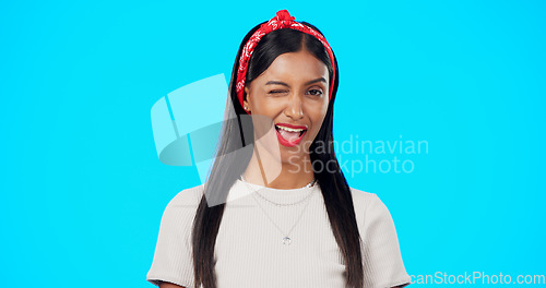 Image of Happy woman, face and wink on blue background, laughing and flirting in studio. Portrait of indian female, model and winking with smile, fun mood and happiness of young gen z fashion, style and joy