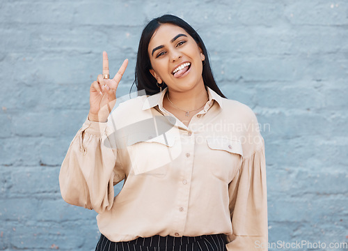 Image of Business woman, portrait and tongue out with peace sign against a gray wall background. Happy and goofy female face smiling showing peaceful hand emoji, sign or gesture with funny or silly expression