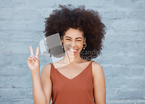 Image of Black woman, portrait and tongue out with peace sign and afro against a gray wall background. Happy and goofy African female face smile showing peaceful hand emoji, sign and funny or silly expression