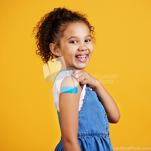 Image of Happy, portrait and child with a plaster in a studio for a wound, sore or injury on her arm. Happiness, smile and healthy girl kid model posing with a bandage after a vaccination by yellow background