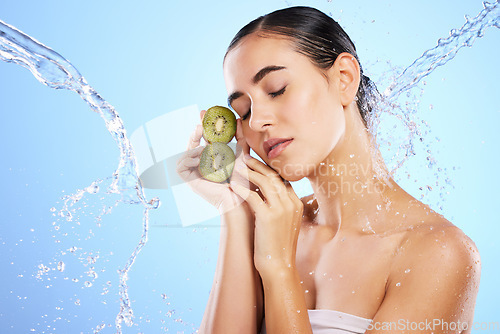 Image of Woman, water splash and face with kiwi in relax for skincare, hygiene or nutrition against blue studio background. Calm or relaxed female holding fruit in natural beauty cosmetics or facial treatment