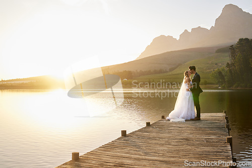 Image of Kiss, sunset and married couple by a lake for a wedding, marriage ceremony and event. Together, happy and a bride and groom standing by a pier, kissing and giving affection to celebrate their love