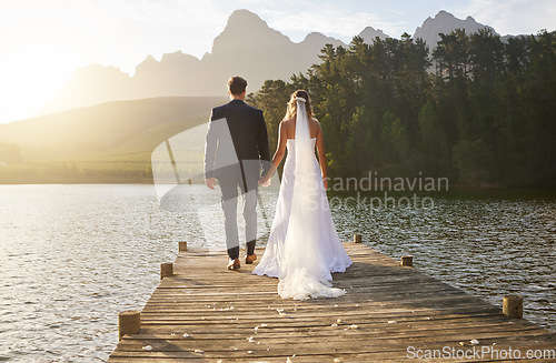 Image of Love, wedding and back of couple by a lake standing, bonding and holding hands on the pier. Nature, romance and young bride and groom in an intimate moment together outdoor on a romantic marriage day