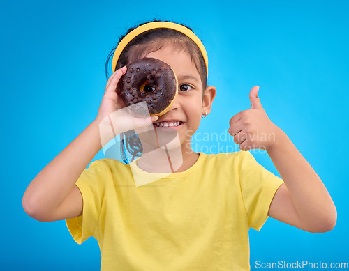 Image of Donut, eye and thumbs up with portrait of girl in studio for junk food, sugar and happiness. Snack, cake and cute with face of child and dessert for eating, positive and chocolate on blue background