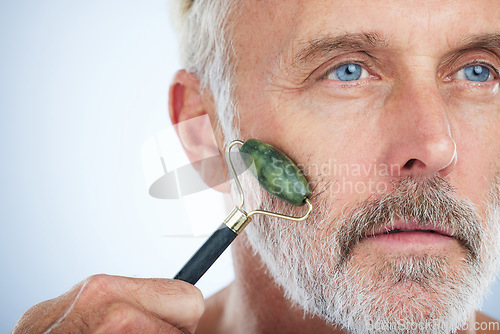 Image of Senior man, jade roller and face for skin with smile, self care and wellness in studio by white background. Elderly model, facial massage and beauty glow with stone product, skincare and dermatology