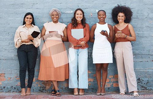 Image of Portrait, group and business women smile on brick wall with devices or gadgets in city. Technology, cooperation and teamwork, diversity or collaboration of friends, people or employees with happiness