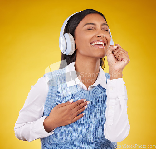 Image of Headphones, happy and woman singing in studio while listening to music, playlist or album. Happiness, smile and Indian female model doing karaoke while streaming a song isolated by yellow background.