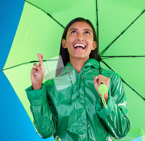 Image of Green, umbrella and fingers crossed with a woman on a blue background in studio during winter for insurance. Rain, fashion or cover and an attractive young female wishing for weather change with hope