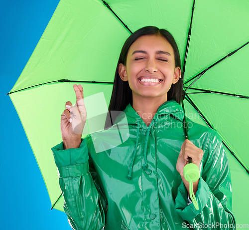 Image of Green, umbrella and fingers crossed with a woman in studio on a blue background during winter for insurance. Rain, fashion or cover and an attractive young female wishing for weather change with hope