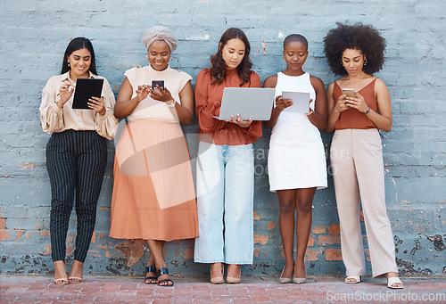 Image of Technology, diversity and business women by a wall standing in a line doing research online. Professional females with phone, tablet and laptop working on a resume while waiting for a job interview.
