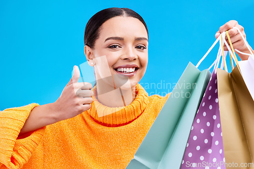 Image of Happy woman, face and shopping bags with thumbs up for purchase, sale or discount against a blue studio background. Portrait of female shopper holding gift bag showing thumb emoji, yes sign or like