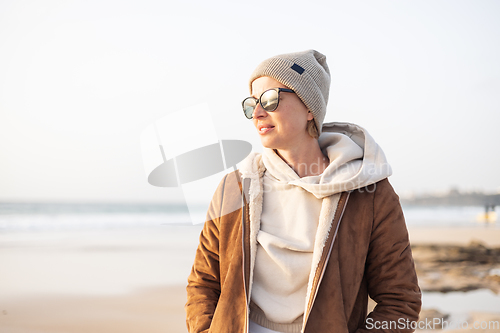 Image of Portrait of young stylish woman wearing brown padded jacket, hoodie, wool cap and sunglasses on long sandy beach in spring.